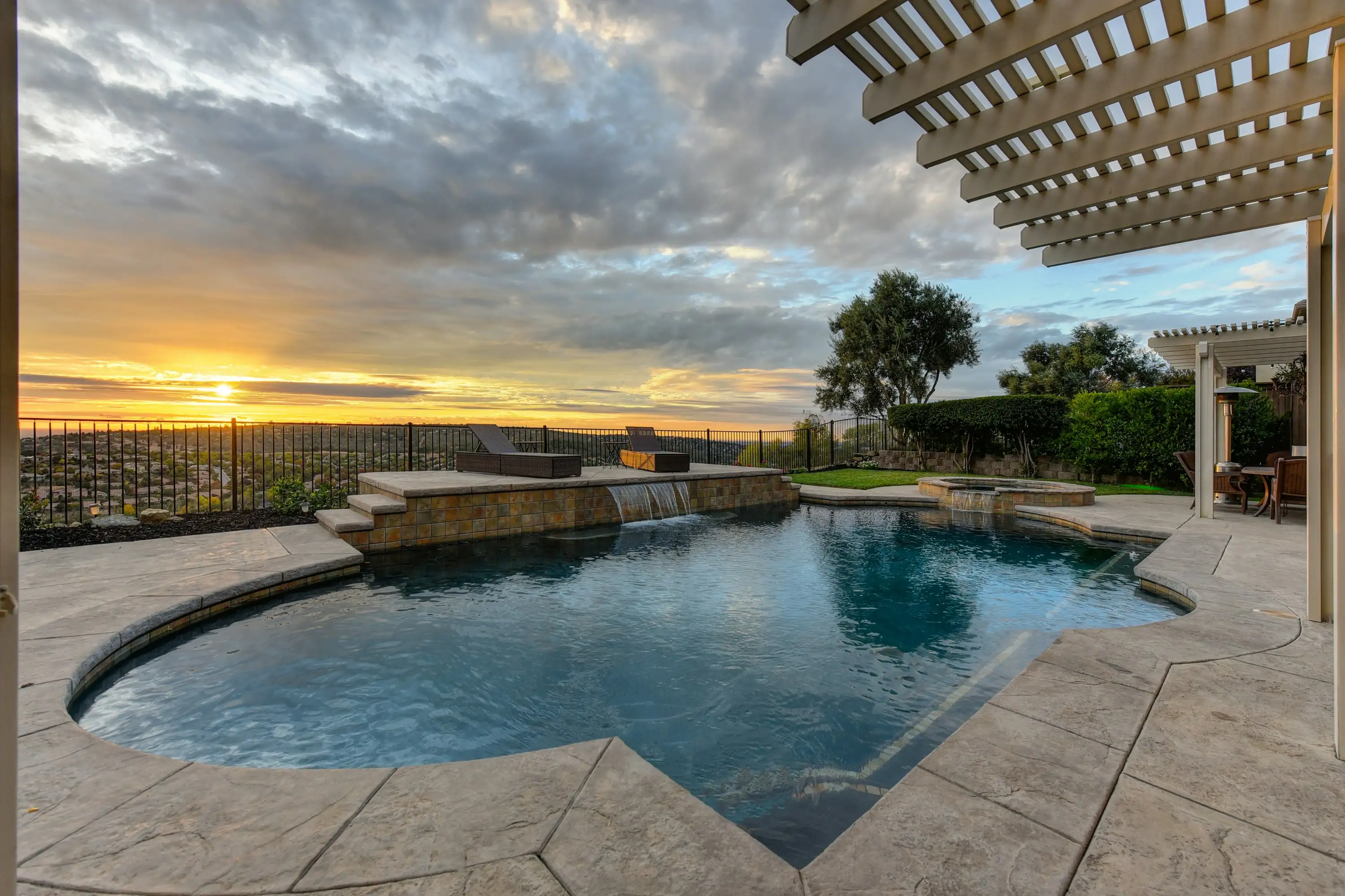 A backyard swimming pool on a flagstone deck at sunset, with open hills beyond the fence